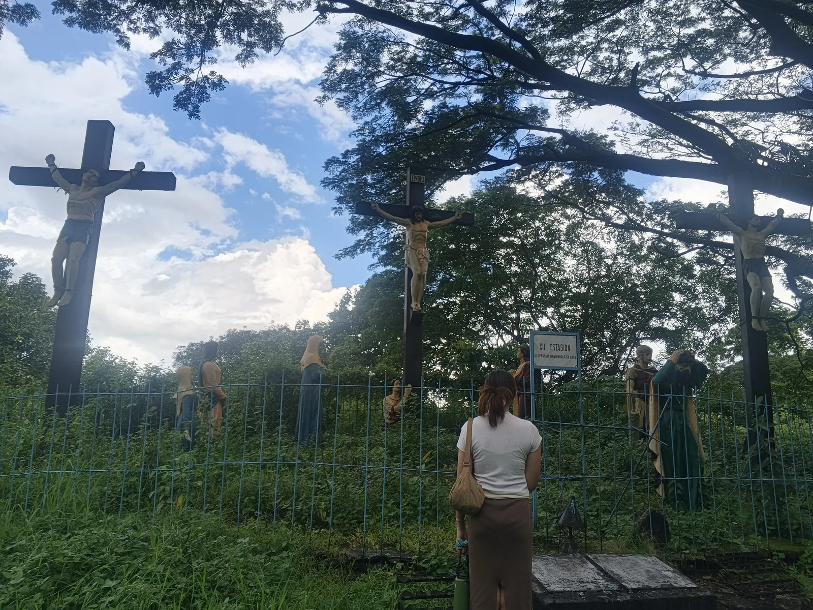 Large cross among trees and devotional figures on the Lourdes Grotto grounds