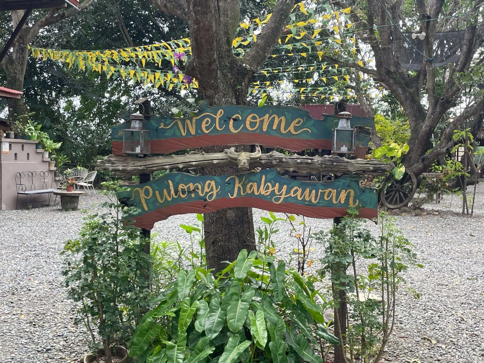 Welcome to Pulong Kabyawan sign amid plants near a tree